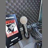Stříbrný dynamický mikrofon s kovovou síťkou a lesklým povrchem těla. Na spodní části mikrofonu je nápis PUTNIK. Vedle něj leží černý box s bílými a…