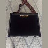 Černý akustický kombozesilovač s texturovaným povrchem a čelní stofovou mřížkou. V horní části je umístěno zlaté logo NUX a pod ním nápis ACOUSTIC. K…