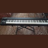 Černý klavírní kontrolér značky Roland, model A-88. Nástroj je umístěn na černém X-stojanu. Na levé straně jsou ovládací prvky, knofíky a tlačítka. P…