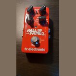 Červený efekt pedál s nápisem HALL OF FAME 2 REVERB a logem tc electronic. Na horní části jsou tři černé otočné ovladače a přepínače s popisky PRE-DE…