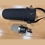 Stříbrný kondenzátorový mikrofon s černým tělem a kovovým držákem. Na těle je viditelné logo značky AKG a nápis R1 ACM-180. Vedle mikrofonu je černý…