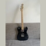 Černá elektrická kytara s černým pickguardem a světlým hnedým hmatníkem. Na hlavě je nápis Fender Mexico. Kytara má dva snímače a tři ovládací knoflí…