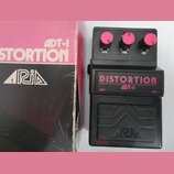 Černý kytarový efekt s nápisem DISTORTION ADT-1 a třemi růžovými oválnými knoflíky s popisky LEVEL, TONE a DIST. Pod efektem je černý plastový kryt s…