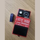 Červený efekt pedál značky BOSS s nápisem Loop Station RC-1. Na horním panelu jsou ovládací prvky LEVEL, tlačítko LOOP a nápis CHECK s STOP/UNDO. Na…