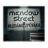 Fotografie zachycuje stříbrný mikrofon na stojanu umístěný před šedou stěnou. Na stěně je černý nápis „mendow street HLEDÁME ZPĚVÁKA“. V pozadí jsou…