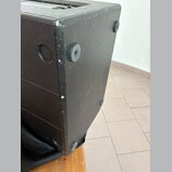 Detail černé reproduktorové bedny s texturovaným povrchem. Na spodní straně jsou vidět čtyři gumové nožičky a na boku kulatý úchop. Hrany bedny vykaz…