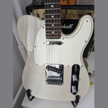 Bílá elektrická kytara typu Telecaster s bílým pickguardem a hnedým hmatníkem. Na pozadí je vidět černý kombozesilovač značky Marshall s nápisem DSL…