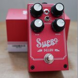 Červený efekt pedál s nápisy Supro a DELAY. Na horní straně jsou čtyři černé ovládací knoflíky s bílými vršekmi a popisky FILTER, REPEATS, LEVEL, TIM…