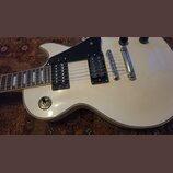 Bílá elektrická kytara s černým pickguardem a dvěma černými humbuckery. Na přepínači tónu jsou viditelné nápisy TREBLE a RHYTHM. Nástroj má chromovan…
