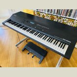 Černý digitální klavír na kovovém stojanu s přiloženým černým pedálem. Na horní části je nápis Roland a vpravo u kláves označení ROLAND DIGITAL PIANO…