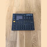 Šedý digitální sampler a sekvencer značky Elektron, model Digitakt. Zařízení má černé ovládací prvky, včetně 16 podsvícených padů a několika otočných…
