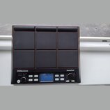 Černý elektronický bicí pad s devíti čtvercovými polštáři v mřížce 3x3. Na spodním panelu jsou nápisy Millenium a NonaPad, malý displej, několik tlač…