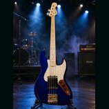Modrá elektrická basová kytara s bílým hmatníkem a bílým perleťovým pickguardem. Na hlavě je nápis Fender Jazz Bass. Nástroj je umístěn na stojanu v…