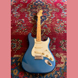 Modrá elektrická kytara typu Stratocaster se světlým javorovým hmatníkem a bílým pickguardem. Na hlavě je vidět nápis Fender Stratocaster. Kytara má…