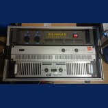 Fotografie zachycuje dva rackové výkonové zesilovače umístěné v transportním kufru. Horní zařízení je černé s nápisem ELMUZ STEREO POWER AMPLIFIER, m…