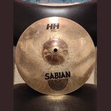 Kovový činel typu splash v zlatavé barvě s viditelným broušením. Na povrchu jsou nápisy HH, Splash 12"/30cm a logo SABIAN s doprovodnými oblouky. Pov…