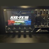 Černý rackový procesor s displejem, na kterém je nápis AXE-FX III PREAMP/FX PROCESSOR MARK II TURBO. V pravém dolním rohu je bílé logo a nápis FRACTA…