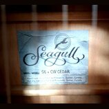 Fotografie znázorňuje štítek uvnitř akustické kytary. Na modrobílém pozadí je černé logo Seagull s vyobrazením ptáka v kruhu. Pod ním je uveden text…