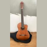 Akustická kytara s vyřezem v korpusu, provedená v přirozené dřevěné barvě. Na hmatníku je vidět štítek s nápisem "Classical Guitar". Nástroj stojí op…