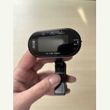 Černý digitální ladicí přístroj s displejem a připevněným klipem. Na levé straně je červené logo AROMA a v dolní části nápis AT-370B TUNER. Horní hra…