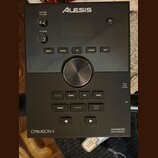 Černý elektronický modul s nápisy ALESIS, CRIMSON II a ADVANCED DRUM MODULE. Zařízení obsahuje několik ovládacích tlačítek s popisky MENU, DOWN, UP,…