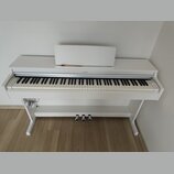 Bílé digitální piano s bílým stojanem a notovým pultem. Na horní liště nad klávesami je umístěno logo YAMAHA. Nástroj má tři st SILVER pedály a někol…