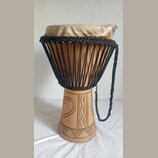 Djembe s tělem z vyřezávaného světlého dřeva a napnutou kůží v horní části. Nástroj je opleten černými provázky, které drží membránu. Spodní část kor…