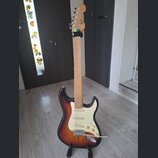 Elektrická kytara typu Stratocaster s javorovým hnedým krkem a tělem v sunburst provedení. Na hlavě krku je svisle napsaný nápis Squier. Kytara má bí…