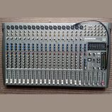 Šedý mixovací pult s nápisy SAMSON, L2000 LIVE MIXING CONSOLE a 24 DSP USB. Zařízení obsahuje řadu vstupních kanálů s modrými a červenými knoflíky, p…