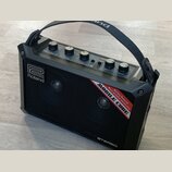 Černý přenosný stereo zesilovač s rukojetí. Na přední mřížce je bílé logo Roland a červenobílý nápis MOBILE CUBE Battery Powered Multi purpose stereo…