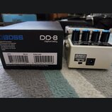 Na fotografii je kytarový efekt Boss DD-8 Digital Delay v bílém provedení s modrými ovládacími knoflíky. Vedle pedálu je umístěna originální černá kr…