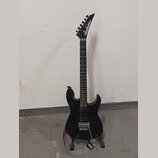 Černá elektrická kytara s špičatou hlavou, na které je bílý nápis Jackson. Nástroj má tmavý hmatník a stříbrný kobylku typu Floyd Rose. Kytara je umí…