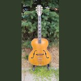 Akustická archtop kytara s přírodním dřevěným povrchem a tmavým hmatníkem. Na hlavě je vidět bílý ornamentální vložkový prvek a šest ladicích kolíčků…