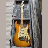 Elektrická kytara s přechodovým žlutě-hnědým lakem v černém tvrdém kufru. Nástroj má bílý pickguard s perleťovým vzorem, tři snímače a javorový hmatn…