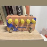 Kytarový efekt pedál v růžovém provedení s žlutými oválnými knoflíky. Na spodní části je modrý nápis phonkiry a pod ním text Tsakalis Audio Works. Na…