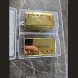 Dvě elektrické kytarové snímače typu humbucker v plastovém průhledném pouzdře. Oba mají zlatý kovový kryt a šest viditelných šroubů. Povrch horního s…