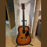 Akustická kytara v barvě sunburst s černým pickguardem a hnědým hmatníkem. Na hlavě je nápis HOHNER. Nástroj stojí na černém stojanu v interiéru. Pov…