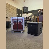 Na fotografii je vidět transportní flightcase v červené barvě s hliníkovými hranami a koly, vedle kterého stojí černý subwoofer. Na flightcasu jsou u…
