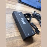 Černý obdélníkový audio rozhraní s nápisy HD 2 a iRig. Zařízení má matný povrch, černý tkaný popruh a vedle něj leží malý plastový černý dílek. Na sp…
