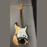 Elektrická kytara typu Stratocaster s černým lakem, který je výrazně odloupaný a odhaluje světlé dřevo korpusu. Na hlavě hmatníku je nápis Fender. Ky…