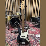 Černá elektrická basová kytara s bílým pickguardem a světle hnědým hmatníkem. Na hlavě je nápis Fender Jazz Bass. Tělo je v lesklém černém provedení,…