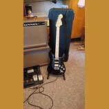 Černá elektrická kytara typu Stratocaster s bílým hmatníkem a bílým pickguardem, opřená o černý tvrdý kufřík. Na hlavě kytary je nápis Fender STRATOC…