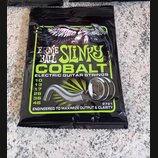 Zavřené balení kytarových strun v černé fólii. Na obalu je nápis ERNIE BALL SLINKY COBALT ELECTRIC GUITAR STRINGS a seznam průměrů 10, 13, 17, 26, 36…