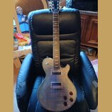 Elektrická kytara typu Les Paul s tmavě hnědým průsvitným lakem a viditnou strukturou dřeva. Nástroj má dva humbuckery, čtyři ovládací knofíky a jede…