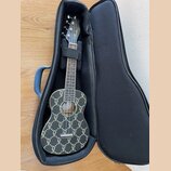 Černý ukulele v černém měkkém pouzdře. Tělo nástroje je pokryto celoplošným zlatým vzorem připomínajícím loga módních značek. Na hlavě je bílý nápis…