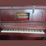 Hnědé dřevěné piano s klaviaturou a dekorativními výřezy na horním panelu. Na levé straně nad klávesami je nápis HUPFELD'S PHONOLISZT a na pravé stra…