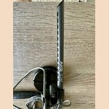 Černý mikrofon s dlouhým, štíhlým trupem a mřížkou v horní části. Na těle je svisle napsán bílý text AKG C747. Mikrofon je umístěn v černém držáku s…
