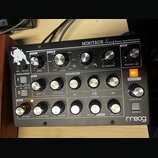 Černý analogový syntetizátor s nápisem MINITAUR analog bass synthesizer a logem Moog v levém dolním rohu. Zařízení má kovové tělo s mnoha stříbrnými…