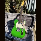 Zelená elektrická kytara typu T-style s bílým pickguardem v otevřeném hrdelním kufru. Na hlavě hmatníku je vidět nápis Fender. Kytara má připevněný v…