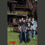 Fotografie zobrazuje skupinu pěti mužů stojících před průmyslovým objektem s kovovými konstrukcemi a žlutými zábranami. V levém dolním rohu je umístě…